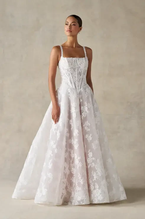 A woman wearing an elegant white wedding gown with floral lace detailing, posing gracefully against a neutral background.