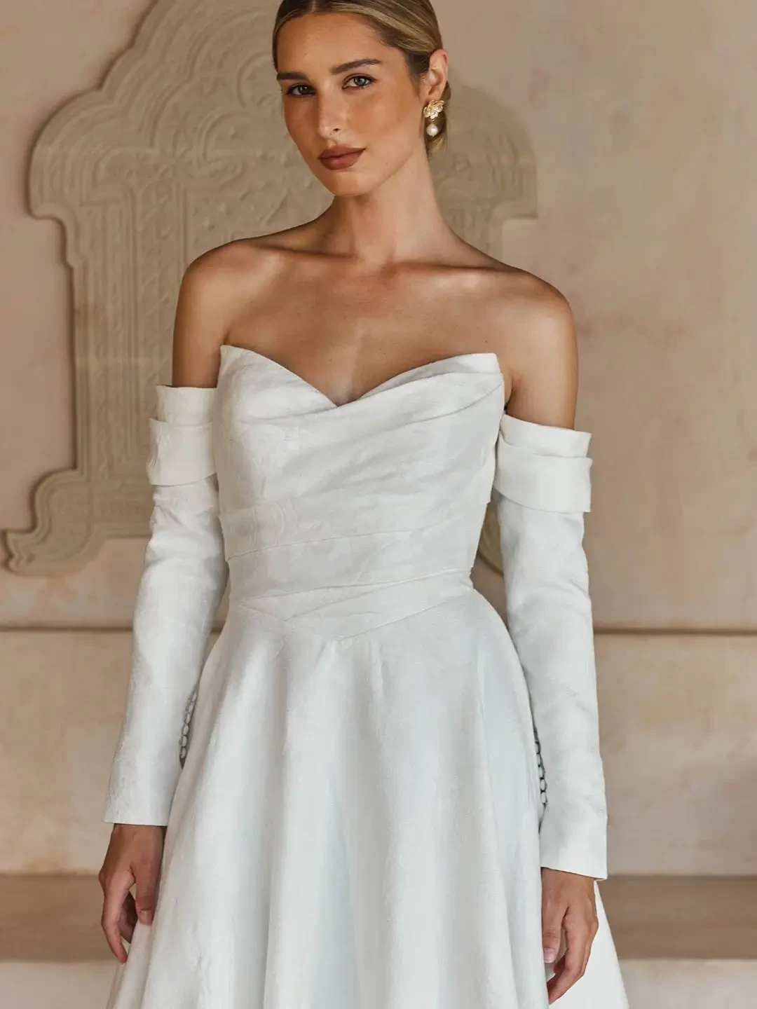 Model is wearing a strapless ivory brocade A-line gown with a sweetheart neckline, Basque waist, and optional statement sleeves and bow.