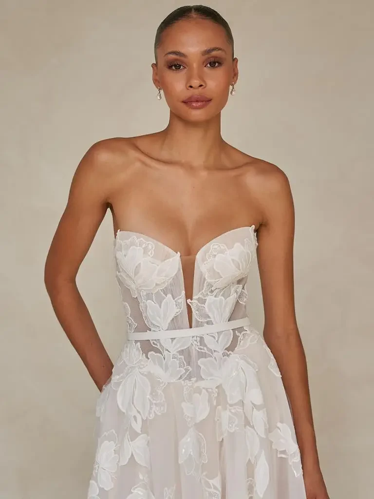 Model is wearing a romantic tulle gown with floral lace detailing, a micro-plunge bodice, and detachable blouson sleeves with ribbon ties.