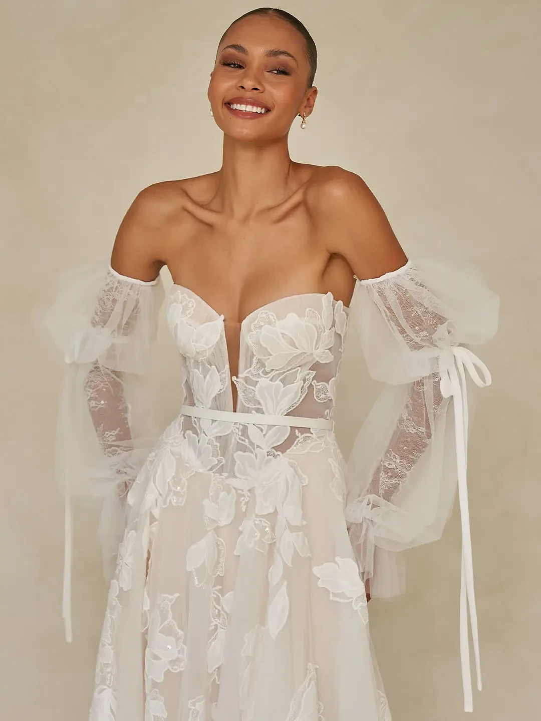 Model is wearing a romantic tulle gown with floral lace detailing, a micro-plunge bodice, and detachable blouson sleeves with ribbon ties.