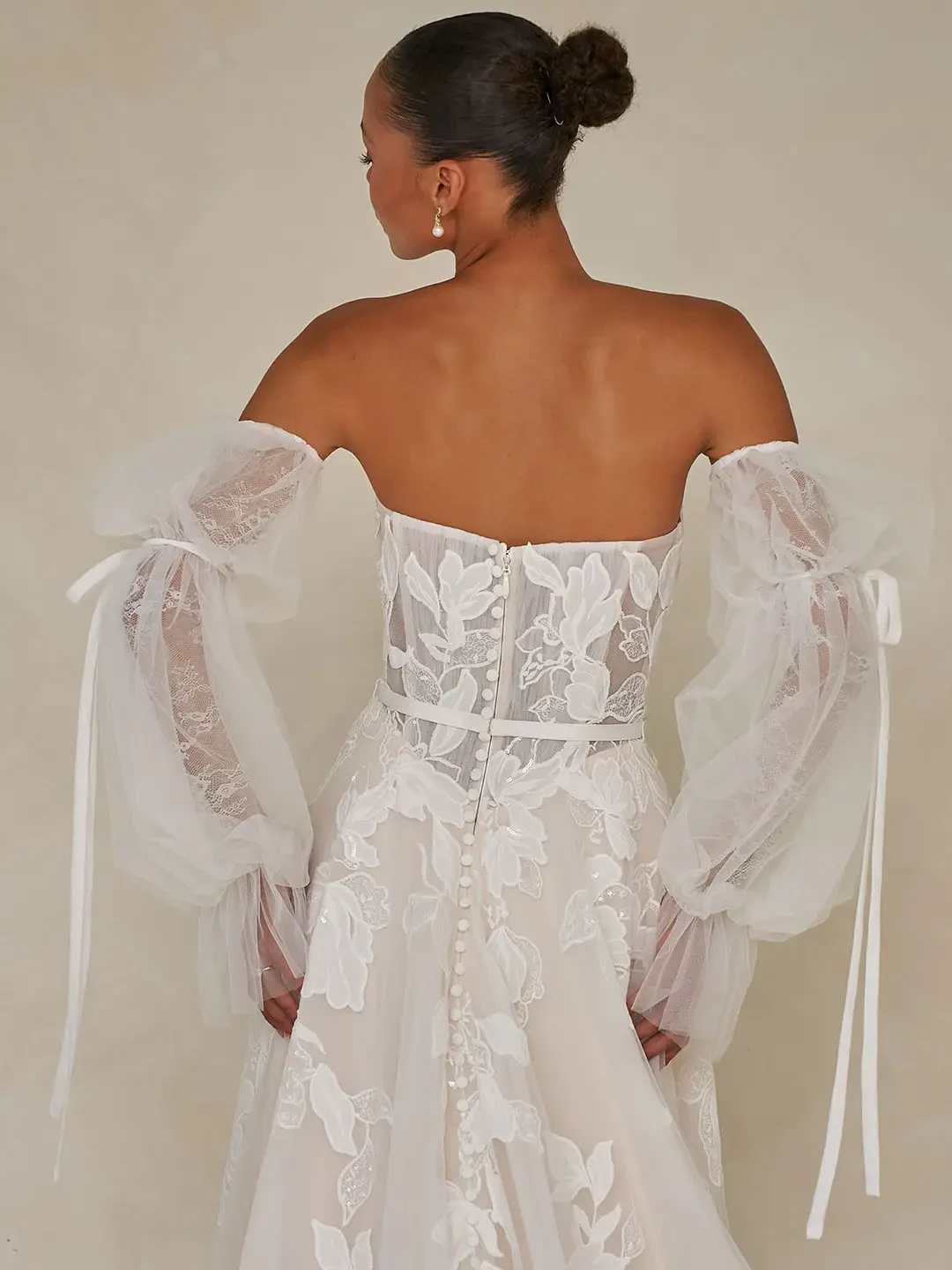 Model is wearing a romantic tulle gown with floral lace detailing, a micro-plunge bodice, and detachable blouson sleeves with ribbon ties.