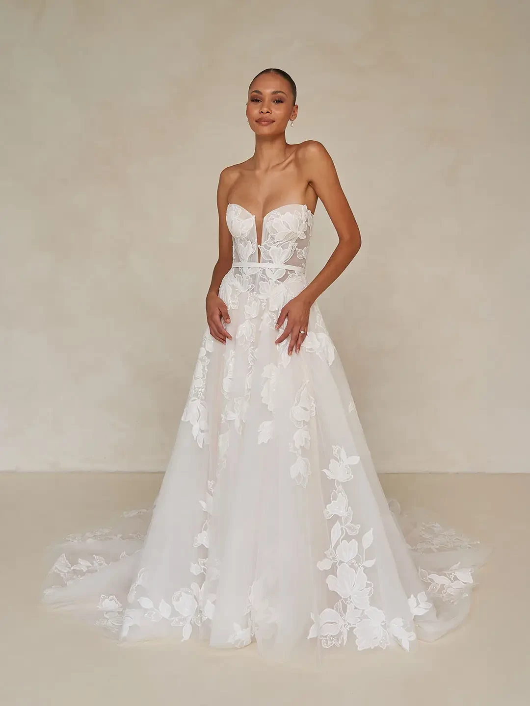 Model is wearing a romantic tulle gown with floral lace detailing, a micro-plunge bodice, and detachable blouson sleeves with ribbon ties.
