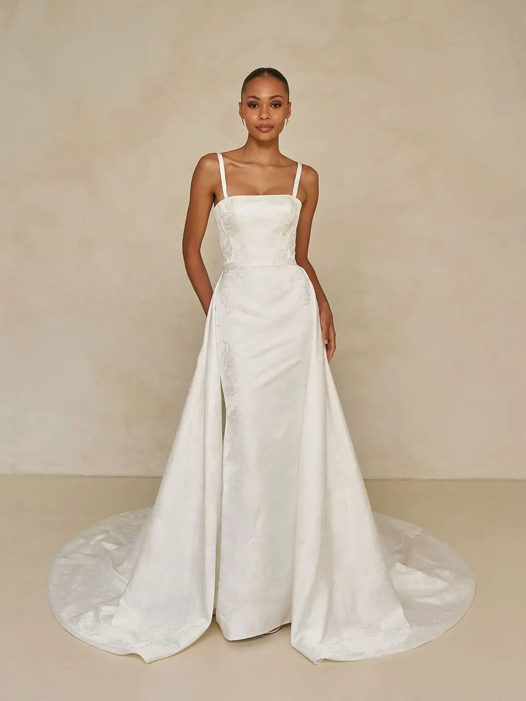Model is wearing a floral jacquard sheath gown with a square neckline, detachable straps, an overskirt for added volume, and an optional back bow.