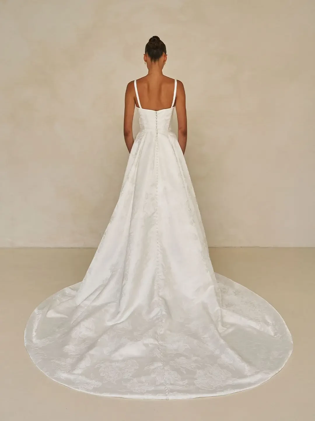 Model is wearing a floral jacquard sheath gown with a square neckline, detachable straps, an overskirt for added volume, and an optional back bow.