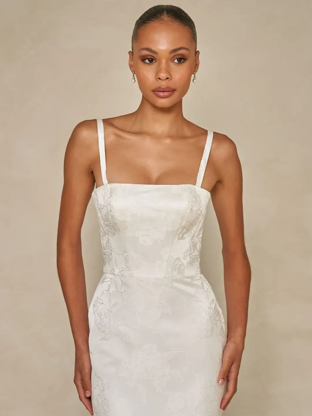 Model is wearing a floral jacquard sheath gown with a square neckline, detachable straps, an overskirt for added volume, and an optional back bow.