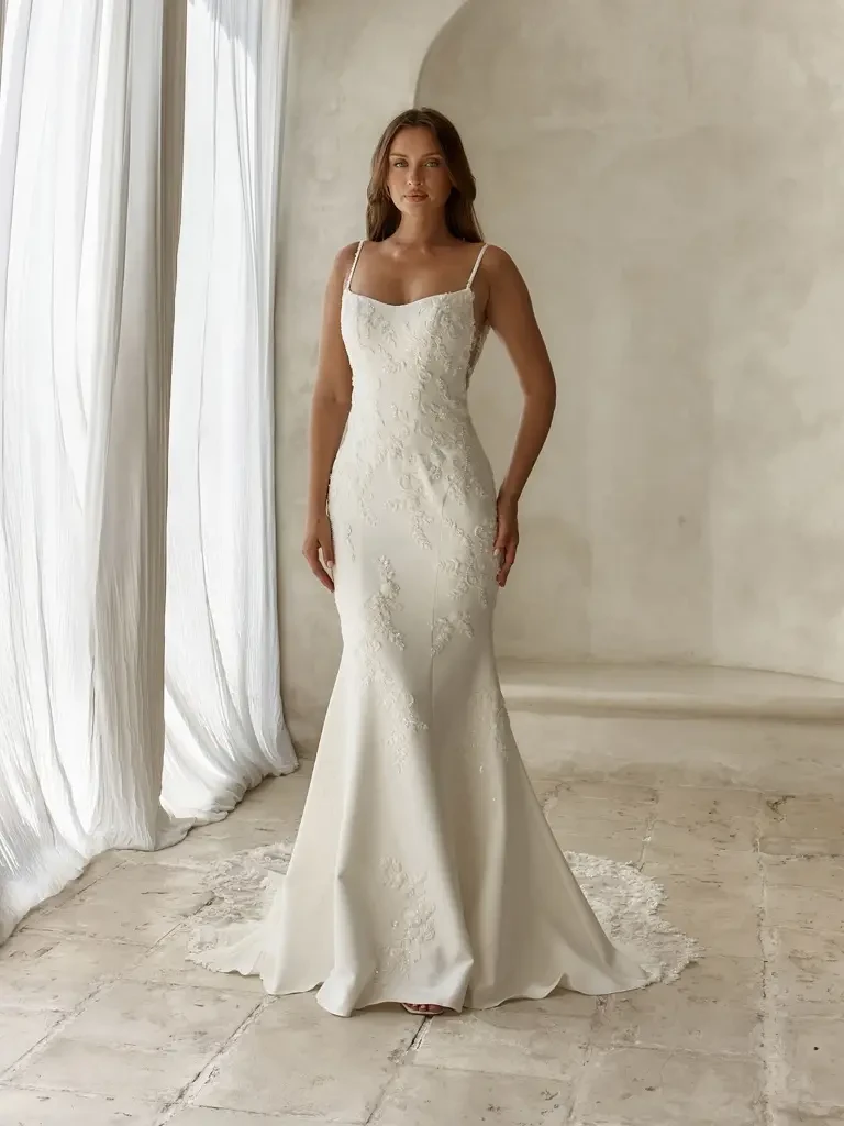 A woman wearing a fitted white wedding dress with a slight train, standing against a neutral background.