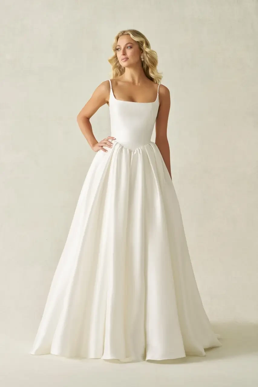 A woman in a simple, elegant white wedding dress with a square neckline and a flowing skirt, standing against a neutral background.