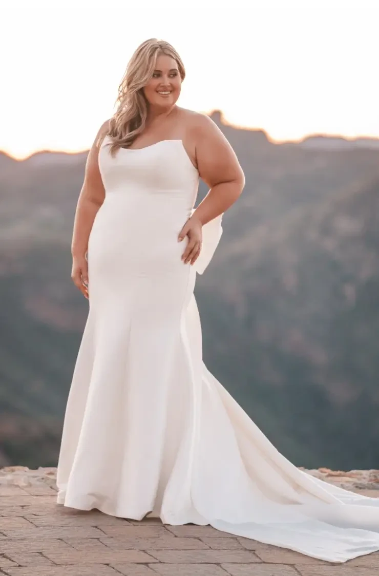 model is wearing a plus size strapless fit and flare minimalist bridal gown with a bow on the back
