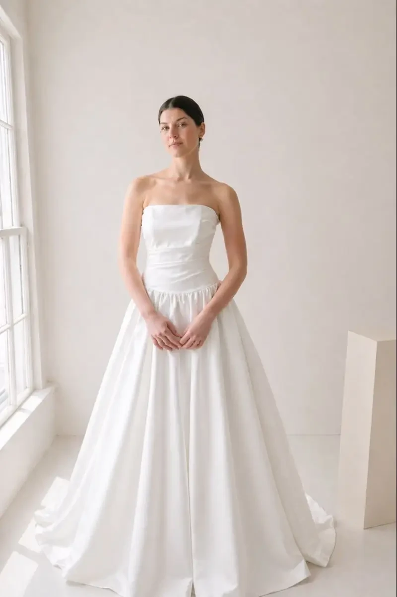 A woman wearing a strapless white wedding dress standing in a bright, minimalistic room.