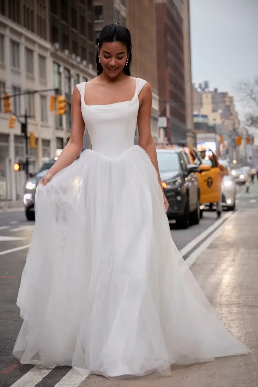 A woman in a white gown stands on a city street, smiling as she looks down, with cars and buildings in the background.