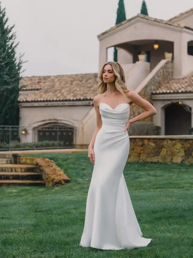 A model wearing a strapless white gown poses outdoors in a garden with a blurred mansion in the background.