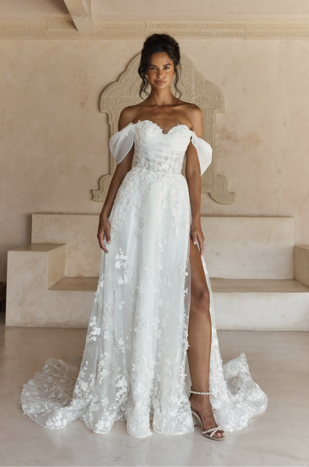 A model wearing a beautiful off-the-shoulder white wedding dress with floral details and a thigh-high slit, standing in a stylish room with neutral tones.