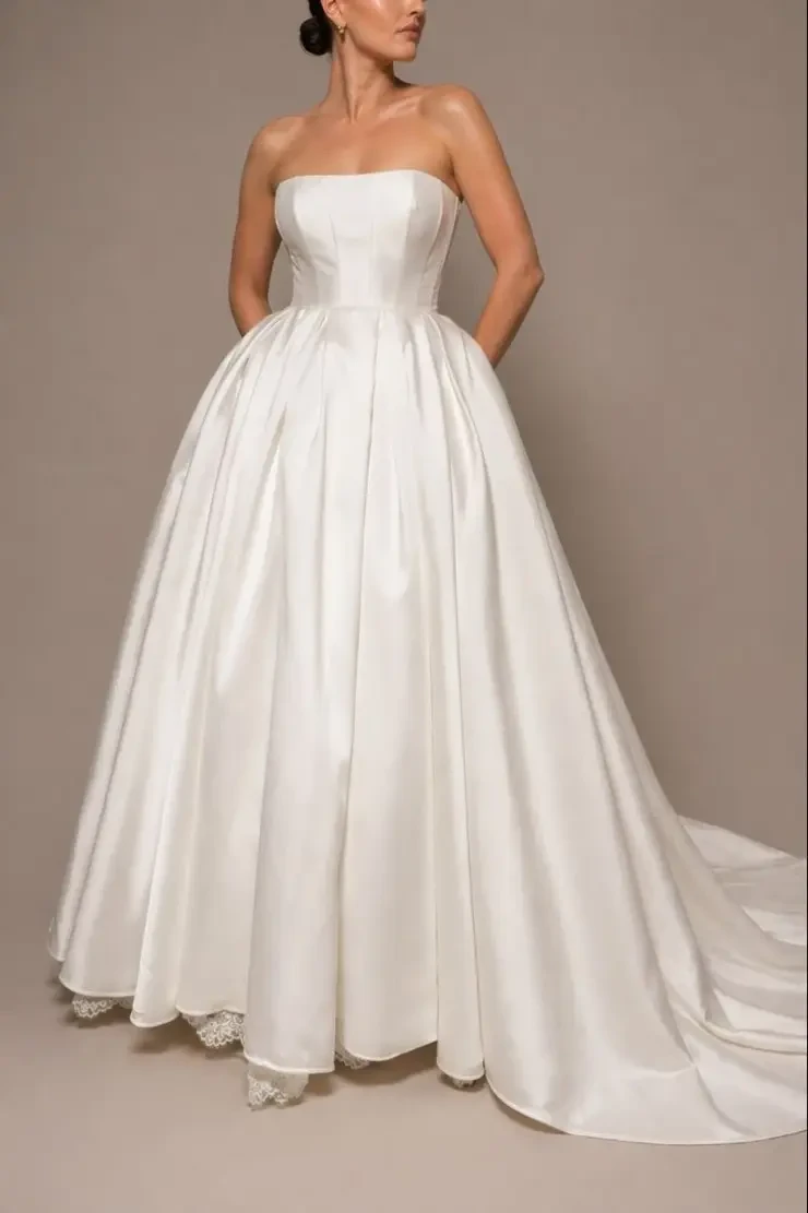 A woman is wearing a strapless, floor-length white gown with a full skirt and pockets.