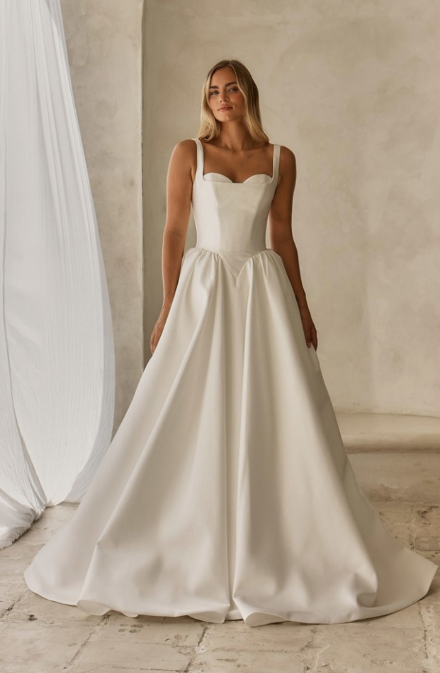 A woman in a white wedding dress with a sweetheart neckline and straps, standing on a light-colored floor against a neutral backdrop.